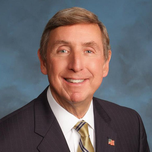 Head shot of a man wearing a suit, gold striped tie, and flag lapel pin in front of a blue background