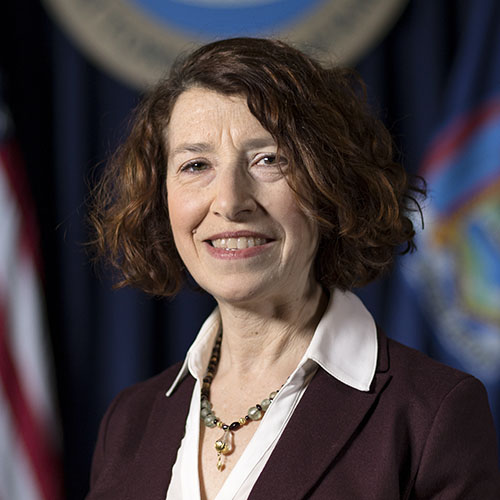 Head shot of a smiling woman in a purple blazer and white collared shirt against various blurry flags