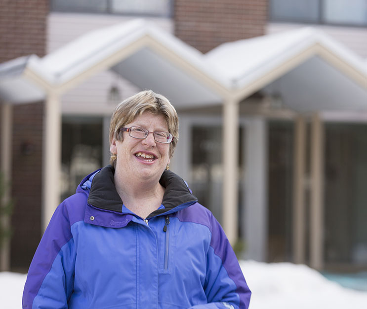 Portrait of a middle-aged woman wearing glasses, smiling, and standing in front of a house