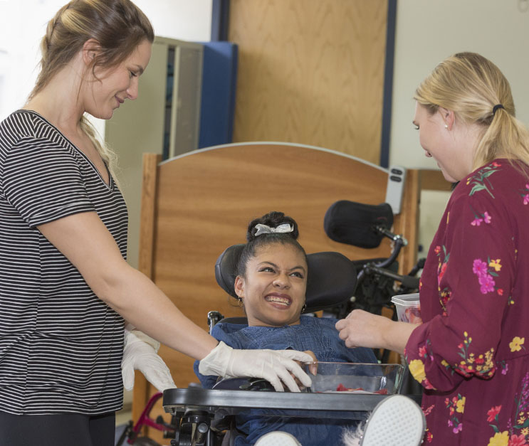 Two female direct support professionals helping woman with a developmental disability seated in a wheelchair
