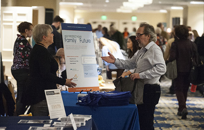 A table of people exhibiting in a convention hallway speak to an attendee on the other side of the table
