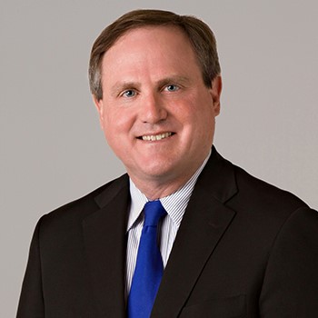 A man in a black suit and blue tie smiles and poses against a gray background for a headshot