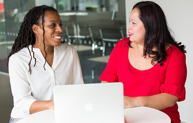 Two women sit at a table looking at each other and smiling with a laptop in front of them
