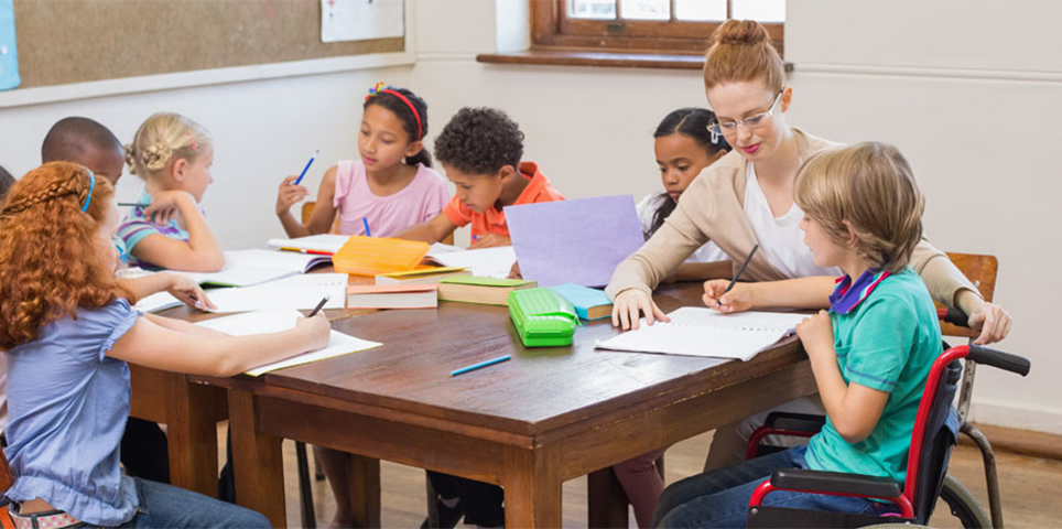 A teacher sits at a table and assists a student as several others around them focus on their work and talk.