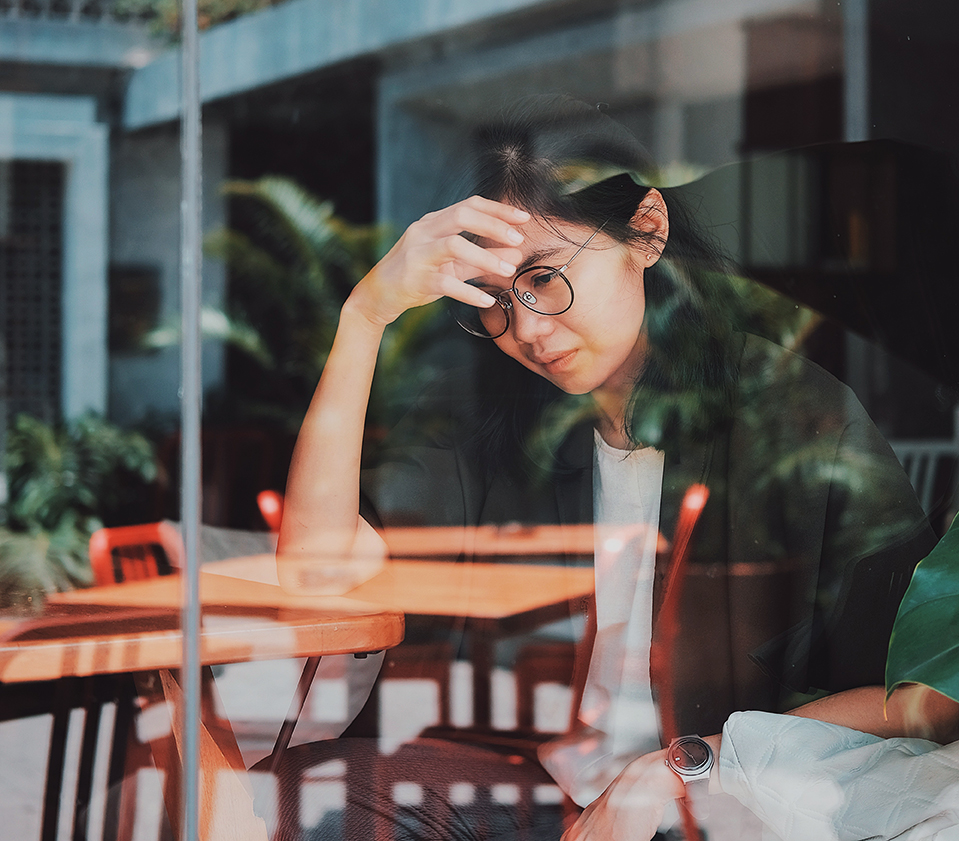 A young woman seen through a window looks upset and has her hand on her head. Outside tables are reflected in the window