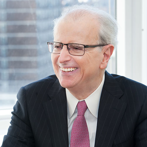 A man smiles, looking away from the camera, and is wearing glasses and a suit with a red tie