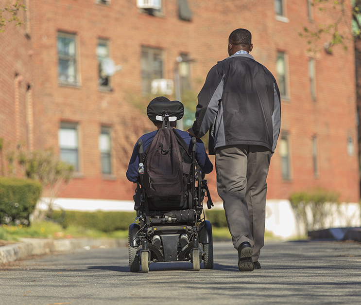 Rear view of a man walking on a paved path alongside another person in a motorized wheelchair. They walk towards a brick building in the background.