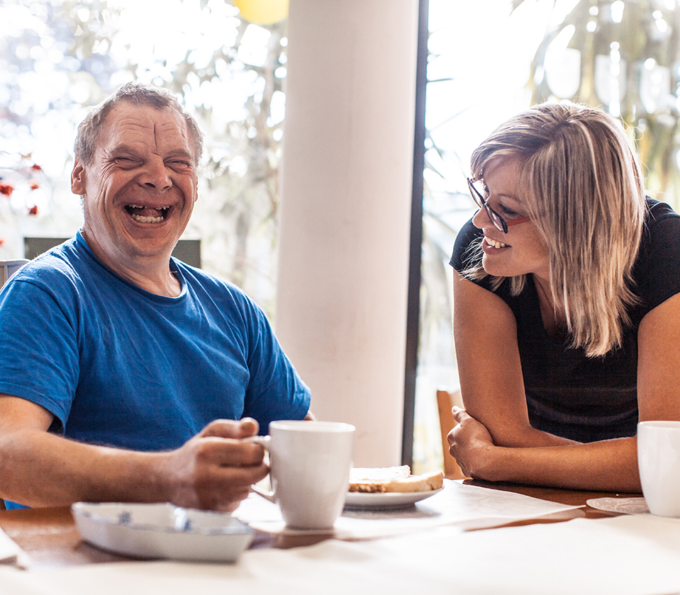 Man smiles as he holds a coffee mug and sits at a table with a woman. She has her arms resting on the table and is smiling at him