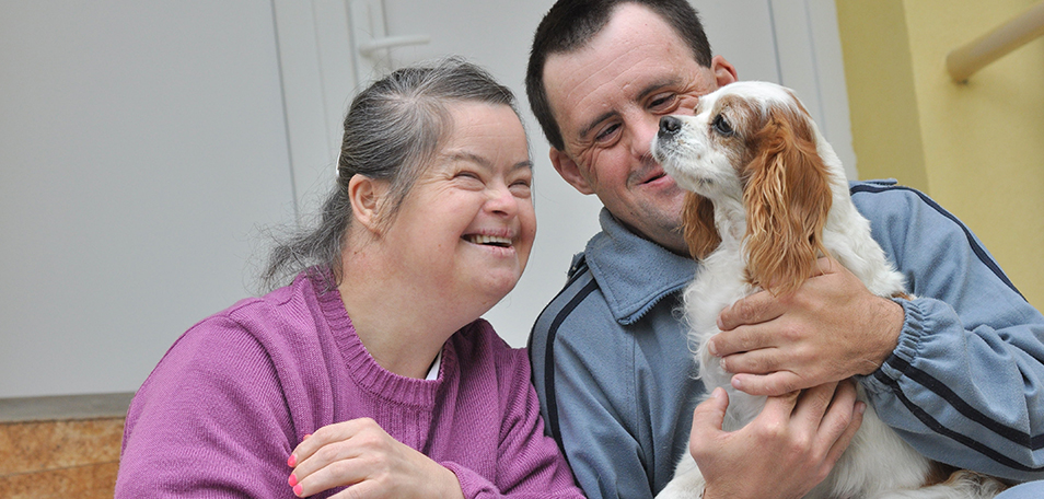 Woman with Down syndrome smiling at a dog that's sitting on the lap of a man sitting beside her.