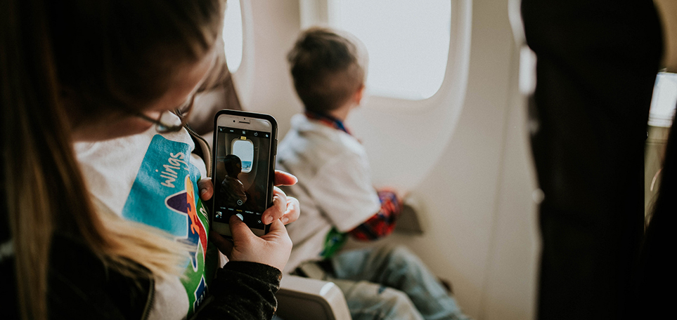 Young girl wearing "Wings" t-shirt on an airplane taking a picture with her cellphone of small boy looking out airplane window